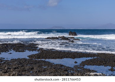 Dynamic turquoise ocean waves splash against dark volcanic rock pools along the scenic Fuerteventura coast. A distant mountain silhouette under a vast blue sky enhances the feeling of peaceful isolation and raw nature.