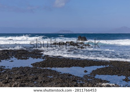 Dynamic turquoise ocean waves splash against dark volcanic rock pools along the scenic Fuerteventura coast. A distant mountain silhouette under a vast blue sky enhances the feeling of peaceful isolation and raw nature.
