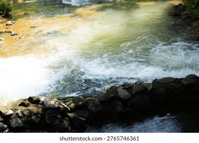 A dynamic view of a fast-flowing stream or small river featuring white water rapids and turbulent currents. The water exhibits shades of green and brown, flowing over rocks and alongside verdant banks, all illuminated by bright natural sunlight creating a shimmering effect.