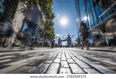 A dynamic view of London's cityscape, featuring Tower Bridge in the distance, with a blurred foreground of a cobblestone street and bustling pedestrians.