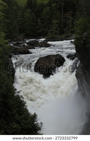 A dynamic view of the rushing rapids at Les Chutes Rawdon in Quebec, Canada. Whitewater cascades forcefully between dark rock walls, surrounded by lush green forest. The natural energy and texture of the fast-moving water highlight the beauty of this popular outdoor destination.