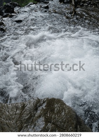 Dynamic white foam forms as rushing water cascades over rocks in a river waterfall, creating a mesmerizing display of movement and texture. The swirling currents and splashing water capture the raw energy of nature while highlighting the freshness and power of flowing freshwater.