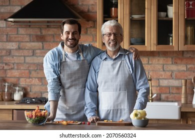 Dynasty Of Chefs. Portrait Of Young Man Grownup Son Hug Hoary Mature Father Look At Camera With Smile Cook Healthy Food At Kitchen. Happy Two Men Of Diverse Family Generations Enjoy Culinary Together