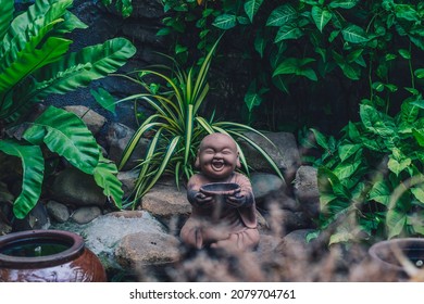 E Statuette figurine symbol brown stone buddha smile hold bowl. Copy space nature tropical leaves plants garden. Mysterious calm soul meditation mood buddhist culture. Bright green, more tone in stock - Powered by Shutterstock