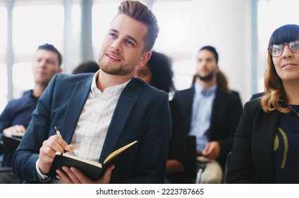 Eager To Hear About This New Brand On The Market. Shot Of A Young Businessman Taking Down Notes While Sitting In The Audience Of A Business Conference.