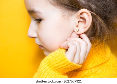 Ear Piercing In A Child - A Girl Shows An Earring In Her Ear Made Of A Medical Alloy. Yellow Background, Portrait Of A Girl In Profile.