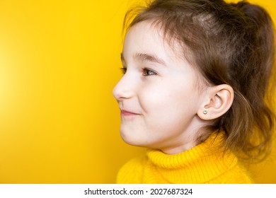 Ear Piercing In A Child - A Girl Shows An Earring In Her Ear Made Of A Medical Alloy. Yellow Background, Portrait Of A Girl In Profile.