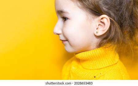 Ear Piercing In A Child - A Girl Shows An Earring In Her Ear Made Of A Medical Alloy. Yellow Background, Portrait Of A Girl In Profile.