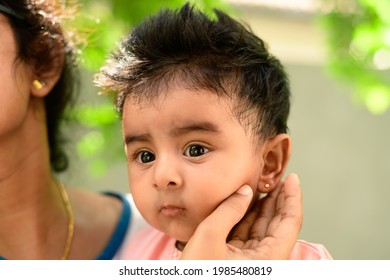 Ear Piercing Day Photoshoot Of A Cute 4 Months Old Baby Girl. Small Gold Earring In Her Ears. Kids Face Close-up As Her Mother Holds The Baby's Ear.