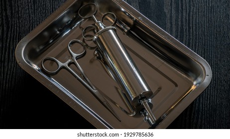 Ear Syringe For Ear Wax Debris Removal And Straight Hemostatic Forceps In A Stainless Steel Container On A Wooden Table Overhead View Close-up Photograph.