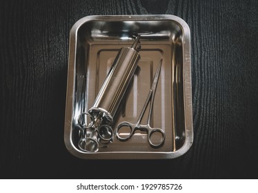 Ear Syringe For Ear Wax Debris Removal And Straight Hemostatic Forceps In A Stainless Steel Container On A Wooden Table Overhead View Close-up Photograph.