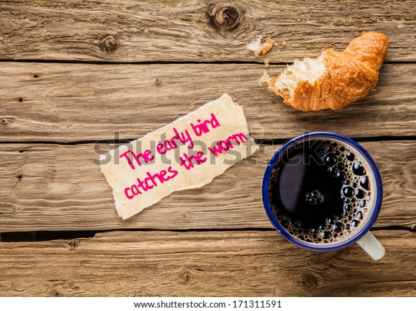 The early bird catches the worm, an\
inspirational saying hand written on a small torn piece of paper\
alongside an early breakfast of frothy espresso coffee and a half\
devoured fresh golden\
croissant