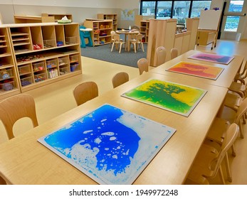 Early Childhood Education Preschool Classroom. Tables With Art, Chairs, And Wooden Shelves With Supplies In Clear Bins. No People. Oil And Water Mix In Squares. 