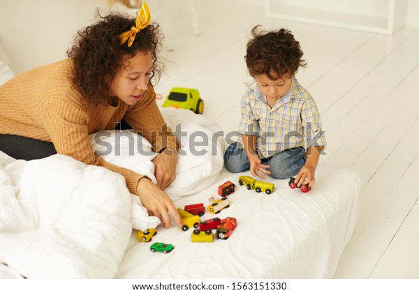 Early development, childhood and maternity. Indoor\
shot of curious cute infant child sitting on floor playing with\
many colorful toy cars on white bed sheet, mother next to him,\
watching him play