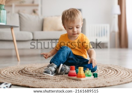 Early development concept. Little toddler boy playing with educational wooden toy at home, sitting in living room interior, free space. Child with wooden colorful stacking and sorting toy