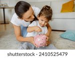 Early financial education. Cute little girl putting coin into pink piggy bank, caring mother teaching her daughter to keep and safe money, sitting on floor in living room at home, closeup