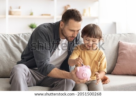 Early financial education. Cute little boy putting coin into pink piggy bank, careful daddy teaching his son to keep and safe money, sitting on couch at home