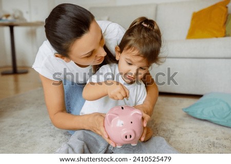 Early financial education. Cute little girl putting coin into pink piggy bank, caring mother teaching her daughter to keep and safe money, sitting on floor in living room at home, closeup