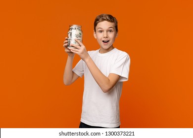 Early Financial Education. Emotional Boy Holding Glass Money Box On Orange Background, Copy Space