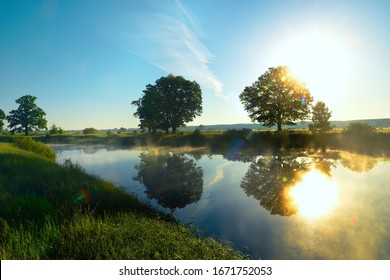 Early morning on the river in the summer, in the spring. the rays of the sun glowing through the trees on the shore. Grass in the dew and fog over the water.
