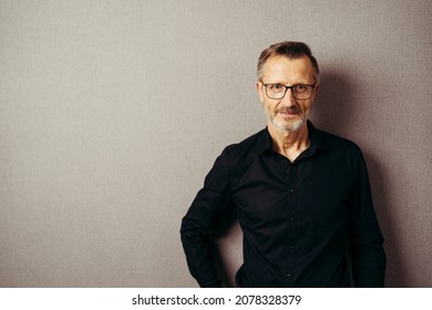 Earnest man wearing glasses in trendy black open necked shirt standing against a beige wall with copyspace looking at camera with a serious expression - Powered by Shutterstock