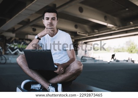 Earnest young athlete with wristwatch in informal wear browsing laptop while sitting with crossed legs under bridge and looking at camera