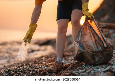 Earth day. Cleanup garbage on the ocean coast. A volunteer with polyethielene bag picking up a plastic bottle on the beach. Close-up. Sunset in the background. The concept of conservation of ecology.