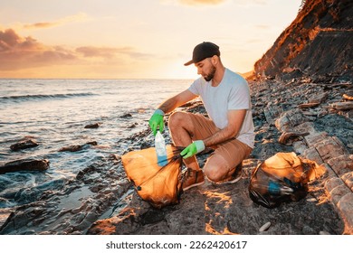 Earth Day. A male volunteer puts a plastic bottle in a bag full of garbage. In the background, the sea and the sunset. Copy space. The concept of environmental conservation. - Powered by Shutterstock