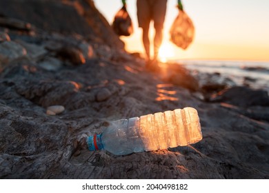 Earth day. Plastic bottle lie at foreground. On the backgroud volunteer carries two large bags of garbage in hands. Sunset on the background. Soft focus. Close up. The concept coastal zone cleaning.