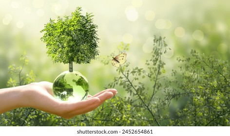 Earth Day or World Environment Day, environmentally friendly concept. Tree growing on globe and yellow butterfly in hand on green background. Save planet and protect nature, sustainable lifestyle.
