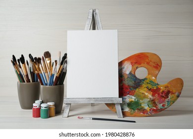 Easel with blank canvas, brushes, paints and palette on white wooden table - Powered by Shutterstock