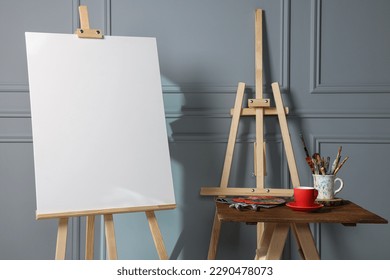 Easel and blank canvas  cup drink   different art supplies wooden table near grey wall