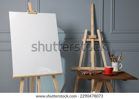 Easel with blank canvas, cup of drink and different art supplies on wooden table near grey wall