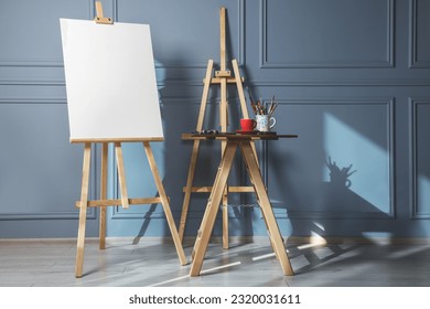 Easel with blank canvas and different art supplies on wooden table near grey wall indoors - Powered by Shutterstock