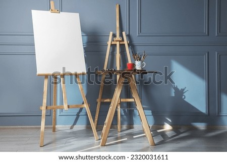 Easel with blank canvas and different art supplies on wooden table near grey wall indoors