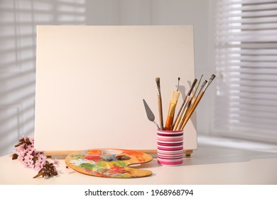 Easel with blank canvas, fresh flowers and art supplies on white table - Powered by Shutterstock