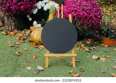 The easel holds a blank canvas surrounded by vibrant fall flowers and pumpkins. Fallen leaves cover the ground, creating a peaceful atmosphere in the garden. - Powered by Shutterstock