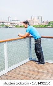 East Indian American Teenager Traveling, Thinking Outside In New York City, Wearing Blue T Shirt, Black Broken Fashionable Jeans, Cloth Shoes, Standing By East River. Brooklyn Buildings On Background.