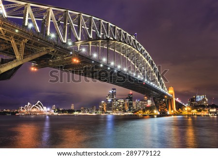 East side of Sydney harbour bridge at sunset with bright illumination of steel arch and columns reflecting in the blurred waters of harbour with Sydney city CBD in the background