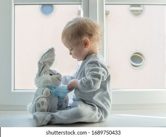 Osterkonzept 2020 mit einem Kind in Hausquarantäne, das im Fenster mit seinem kranken, plüschigen Hasen spielt und eine medizinische Maske gegen Viren während des Coronavirus COVID-2019 und Grippe-Ausbruchs trägt.