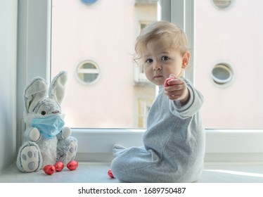 Osterkonzept 2020 mit einem Kind in Hausquarantäne, das im Fenster mit seinem kranken, plüschigen Hasen spielt und eine medizinische Maske gegen Viren während des Coronavirus COVID-2019 und Grippe-Ausbruchs trägt.