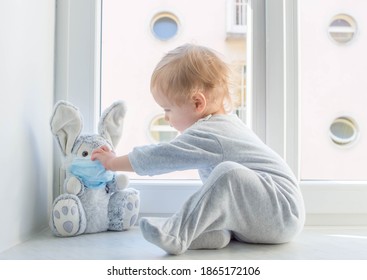 Osterkonzept 2021 mit einem Kind in Hausquarantäne, das im Fenster mit seinem kranken, plüschigen Hasen spielt, der eine medizinische Maske gegen Viren während des Coronavirus COVID-2019 trägt und die Grippe abschließt.