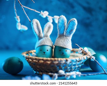 Easter blue eggs in crocheted hats with bunny ears. Eggs in a basket, willow branch, blue background