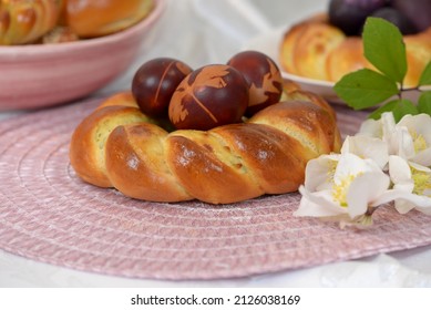 Easter bread wreath with Easter eggs on pink table napkin with hellebores. Pink bowl  with more home baked bread rolls and plate with braided bread and home coloured Easter eggs in background.