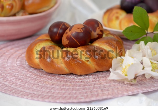 Easter\
bread wreath with Easter eggs on pink table napkin with hellebores.\
Pink bowl  with more home baked bread rolls and plate with braided\
bread and home coloured Easter eggs in\
background.