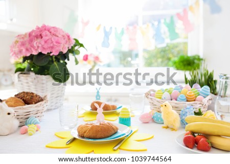 Easter breakfast table setting. Decoration for Easter family celebration. Eggs basket and spring flowers. Bread, croissant and fruit for kids meal. Egg and pastel bunny decor in kitchen at window.