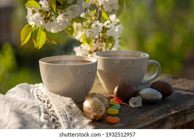 Easter Brunch Outdoor. Coffee, Cocoa Drink, Hot Chocolate, Tea With Milk In Cups With Heart Decor And Colorful Eggs, Bean-shaped Lollipops, Blossoming Cherry Branches On A Wooden Table.