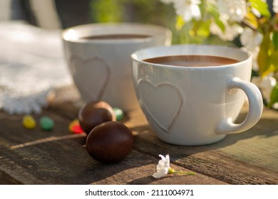 Easter Brunch Outdoor. Coffee, Cocoa Drink, Hot Chocolate, Tea With Milk In Cups With Heart Decor And Colorful Eggs, Bean-shaped Lollipops, Blossoming Cherry Branches On A Wooden Table