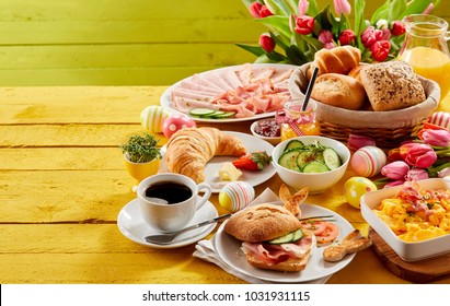 Easter buffet breakfast or brunch with assorted bread rolls, cheese, meat, scrambled egg, orange juice, coffee on a wooden table decorated with easter eggs and spring flowers with copy space