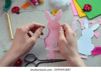 Easter Bunny Felt Sewing Tutorial in Progress. Hands Stitching Pink Rabbit Shape with Needle, Surrounded by Colorful Materials, Flowers, Ladybugs, Threads, and Craft Tools for DIY Decoration